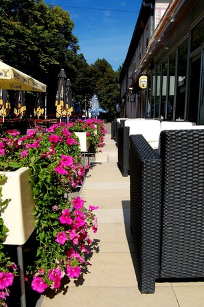 A row of outdoor seating with pink flowers, black wicker furniture, and a building with a sign in the background. - Tiski Cvet Hotel (Novi Becej)