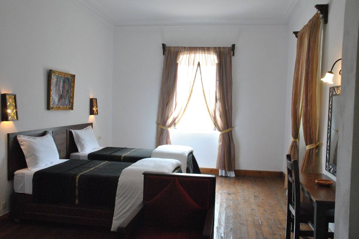 A hotel room with two beds, a wooden floor, a painting on the wall, and a window with curtains.