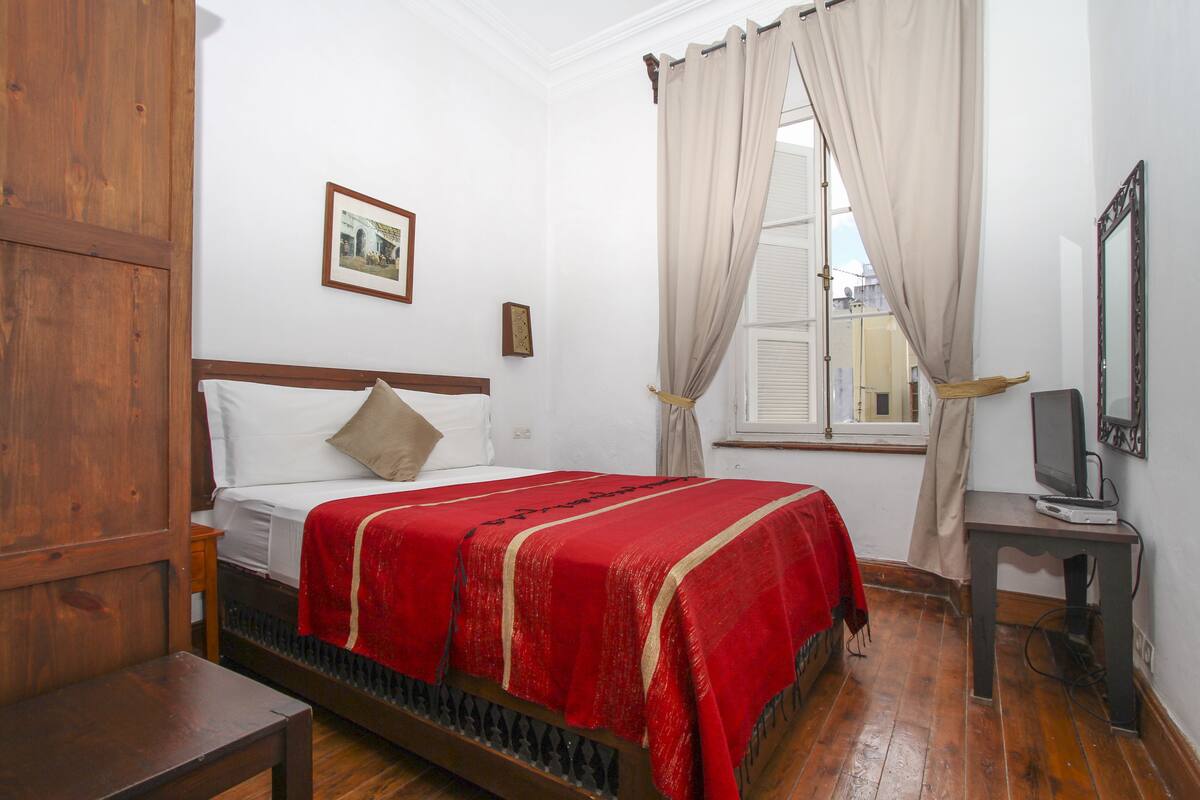 A bedroom with a wooden bed, a red blanket, a wooden nightstand, a television, and a window with curtains.
