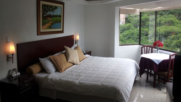 Habitación de hotel con una cama grande, un cuadro en la pared, un ventanal con vistas a la vegetación y una mesita con un jarrón de flores.