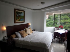 A hotel room with a large bed, a painting on the wall, a window with a view of greenery, and a small table with a vase of flowers. - Hotel Plaza Mayor Cali (Cali)