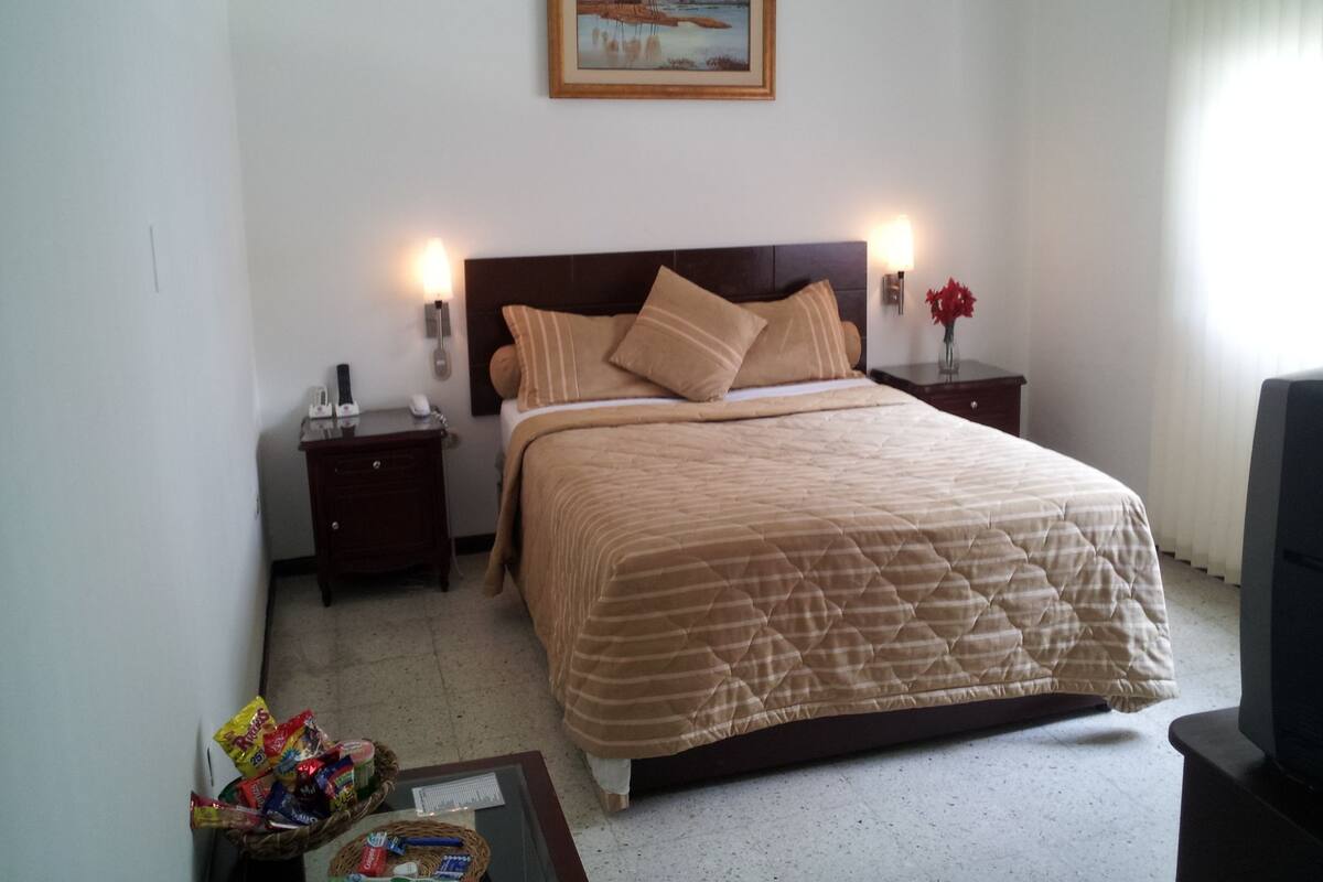 A neatly arranged hotel room with a large bed, two bedside tables, a nightstand, a small table with snacks, and a framed picture on the wall.