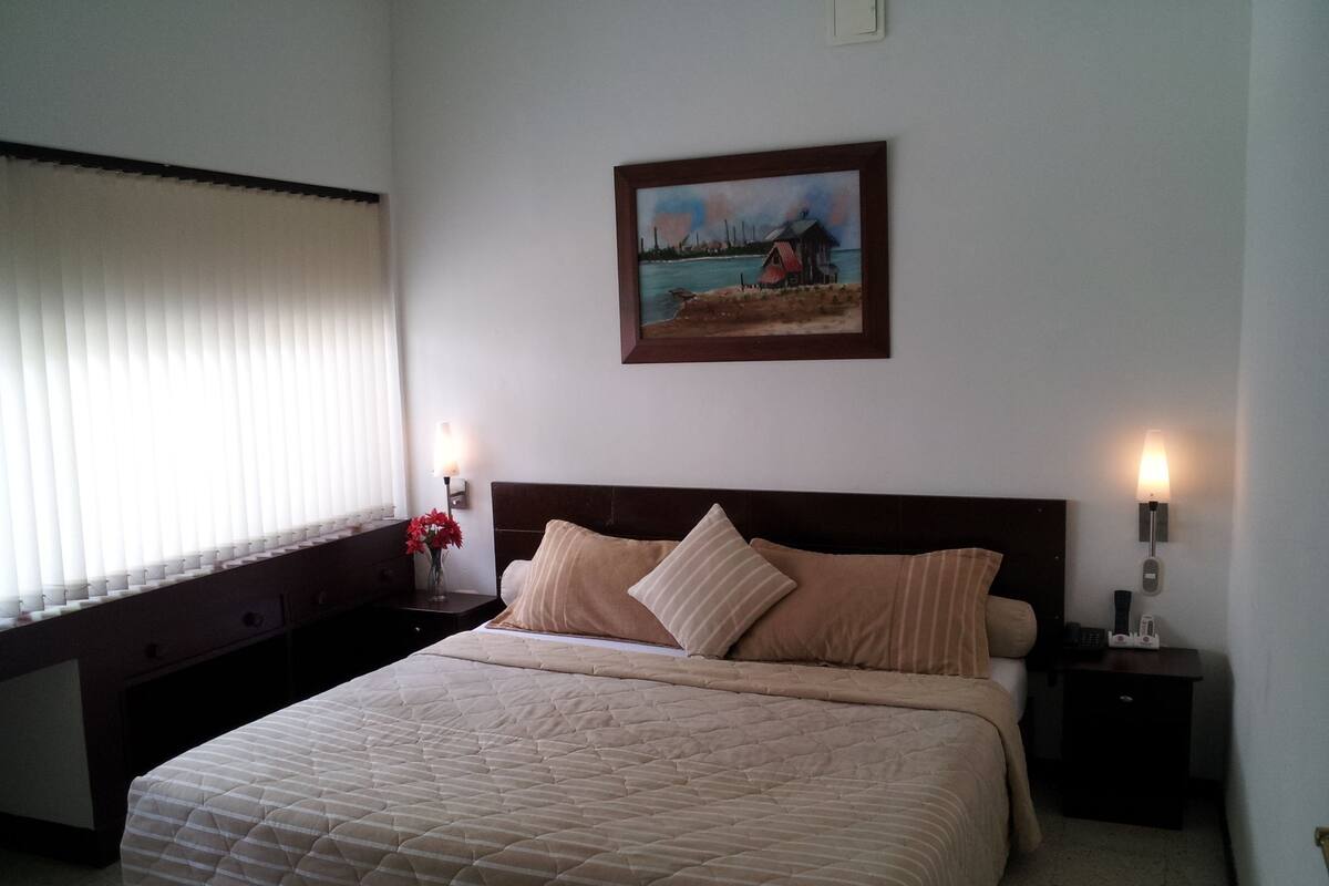 A bedroom with a large bed, a nightstand with a lamp, and a painting on the wall.