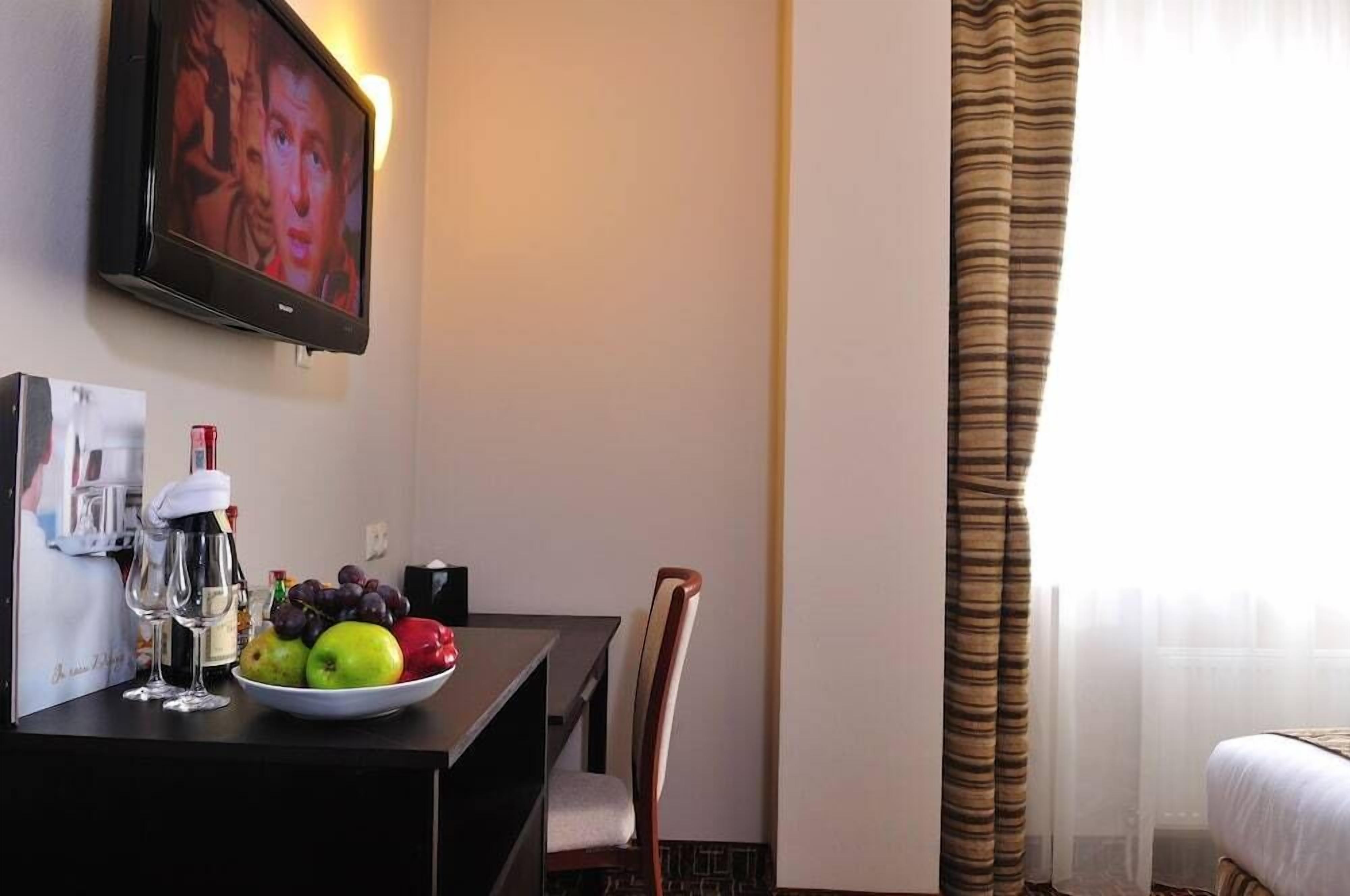 A hotel room with a bed, a TV mounted on the wall, a dining table with a bowl of fruit and glasses, and a chair.