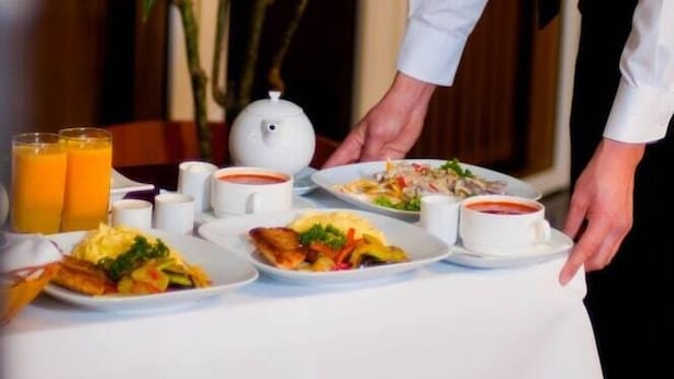 A waiter serving breakfast in a room with a large plant and a white tablecloth.