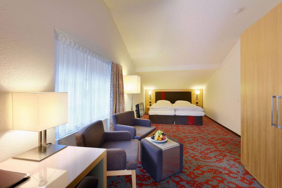 A hotel room with a bed, two armchairs, a small table with a fruit bowl, a desk with a lamp, and a wardrobe.