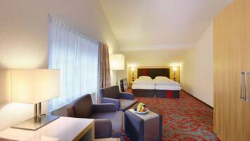 A hotel room with a bed, two armchairs, a small table with a fruit bowl, a desk with a lamp, and a wardrobe.