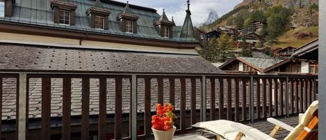 A wooden deck with lounge chairs and a flower pot, overlooking a mountain village with traditional houses and a steep hillside.