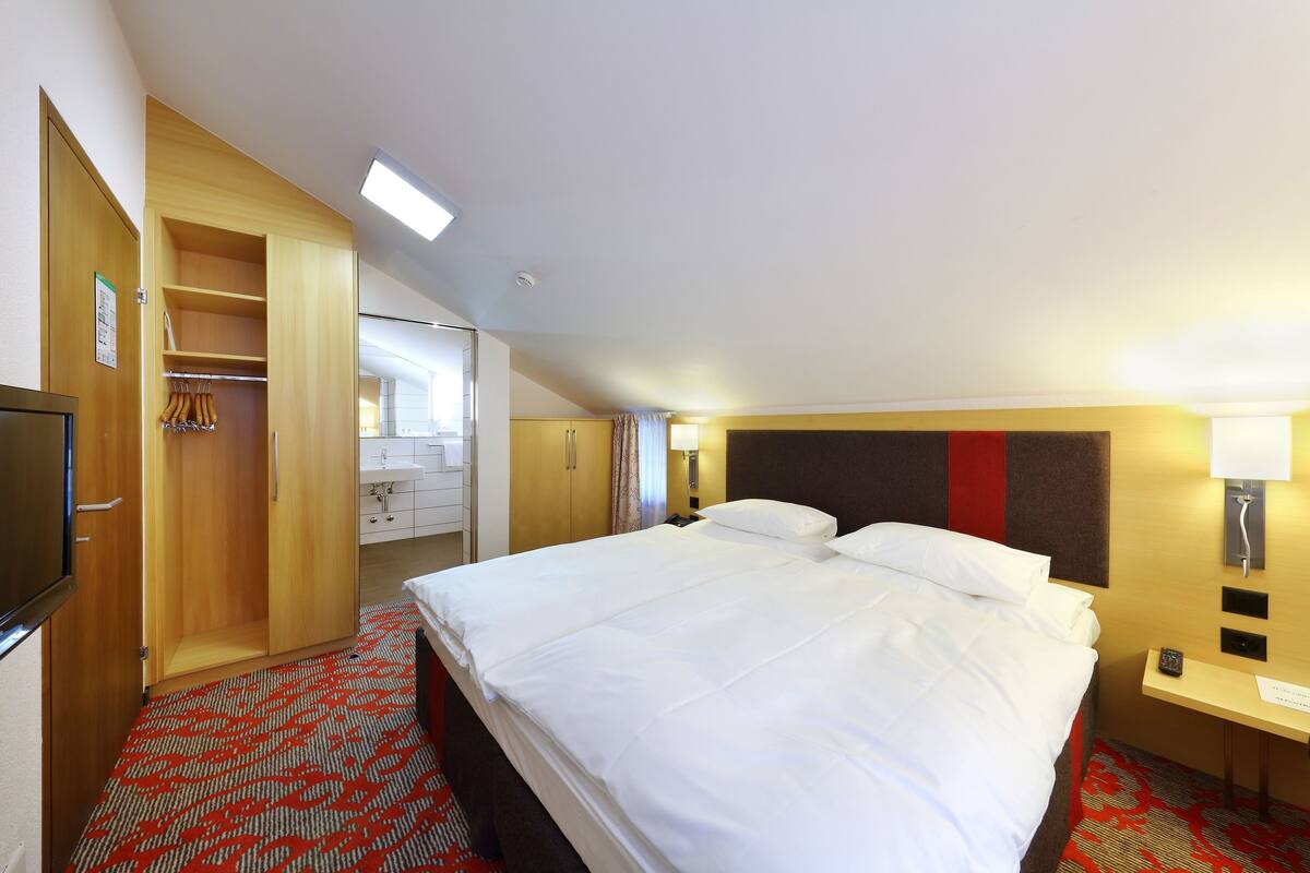 A hotel room with a large bed, a wardrobe, a desk with a lamp, and a bathroom visible through an open door.
