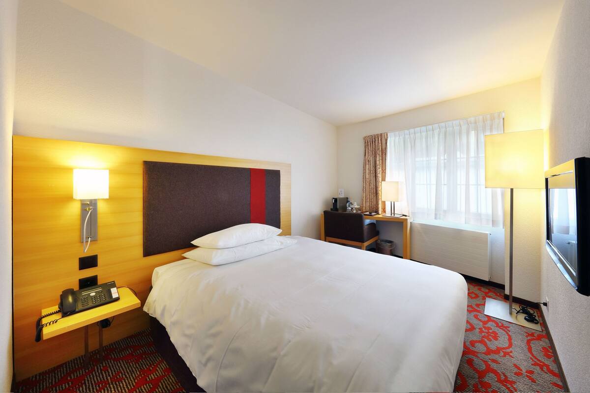 A hotel room with a bed, bedside table with a phone, a desk with a lamp, and a TV on the wall.