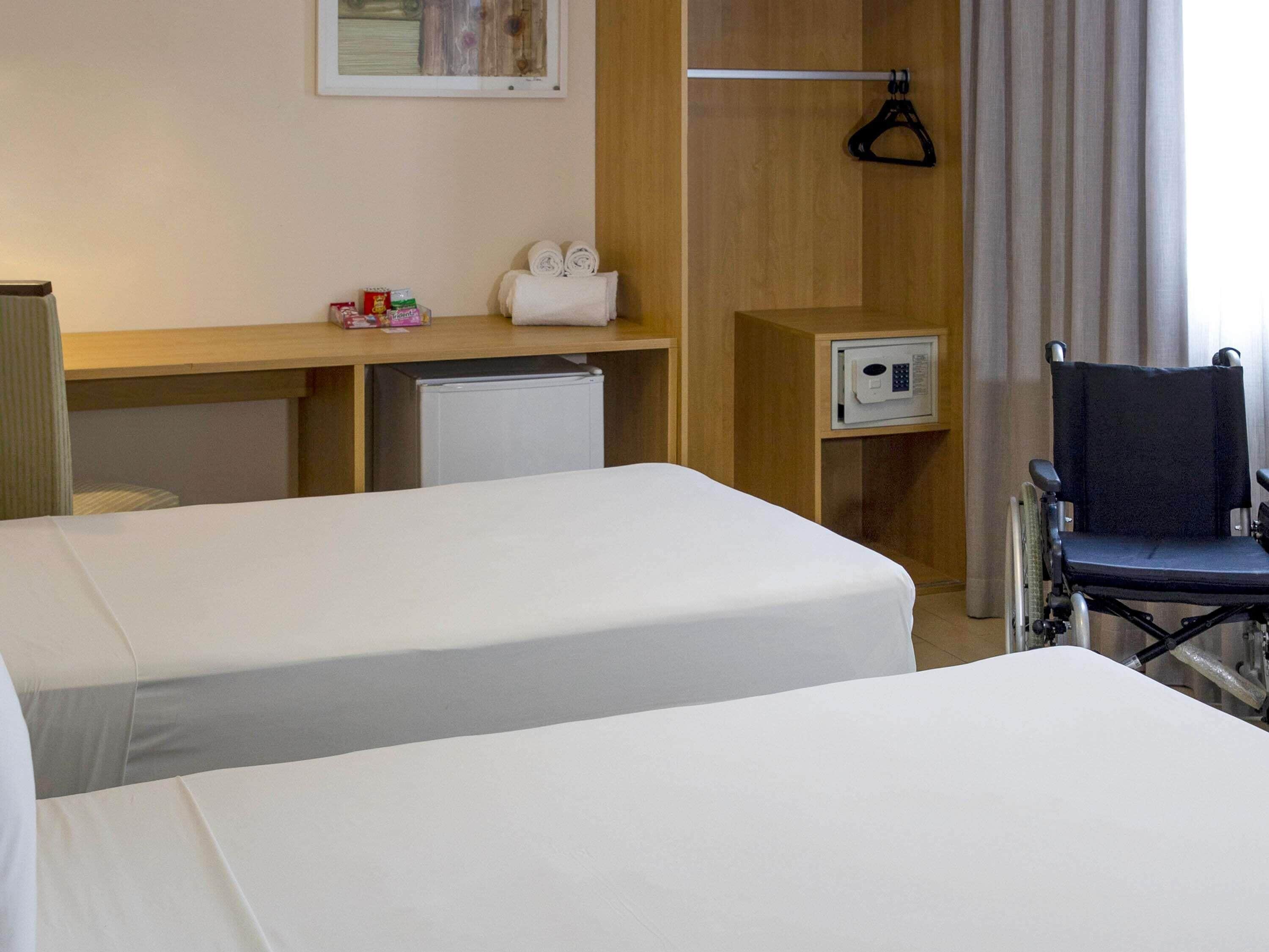 A hotel room with two beds, a desk, a microwave, a wheelchair, and a hanging coat rack.