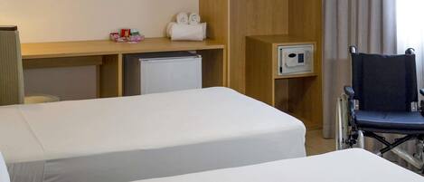 A hotel room with two beds, a desk, a microwave, a wheelchair, and a hanging coat rack.
