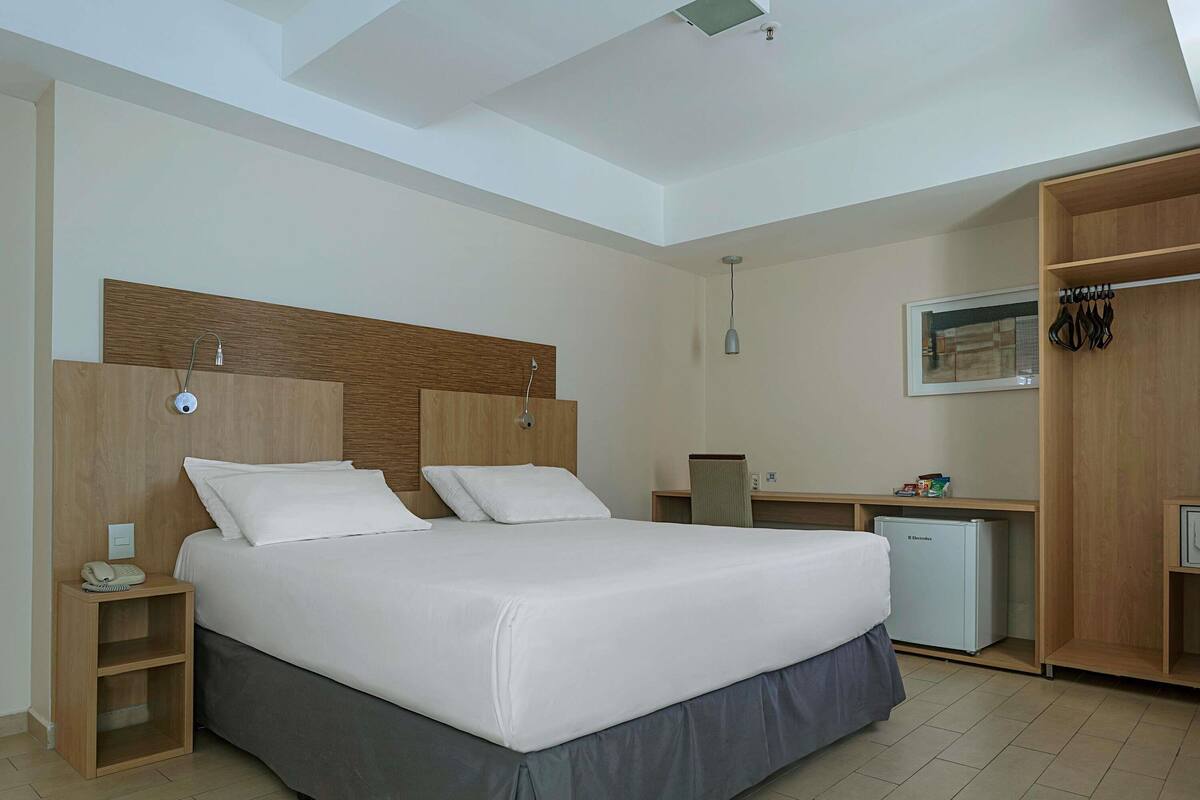 A hotel room with a bed, bedside table, desk, chair, wardrobe, and a small refrigerator.