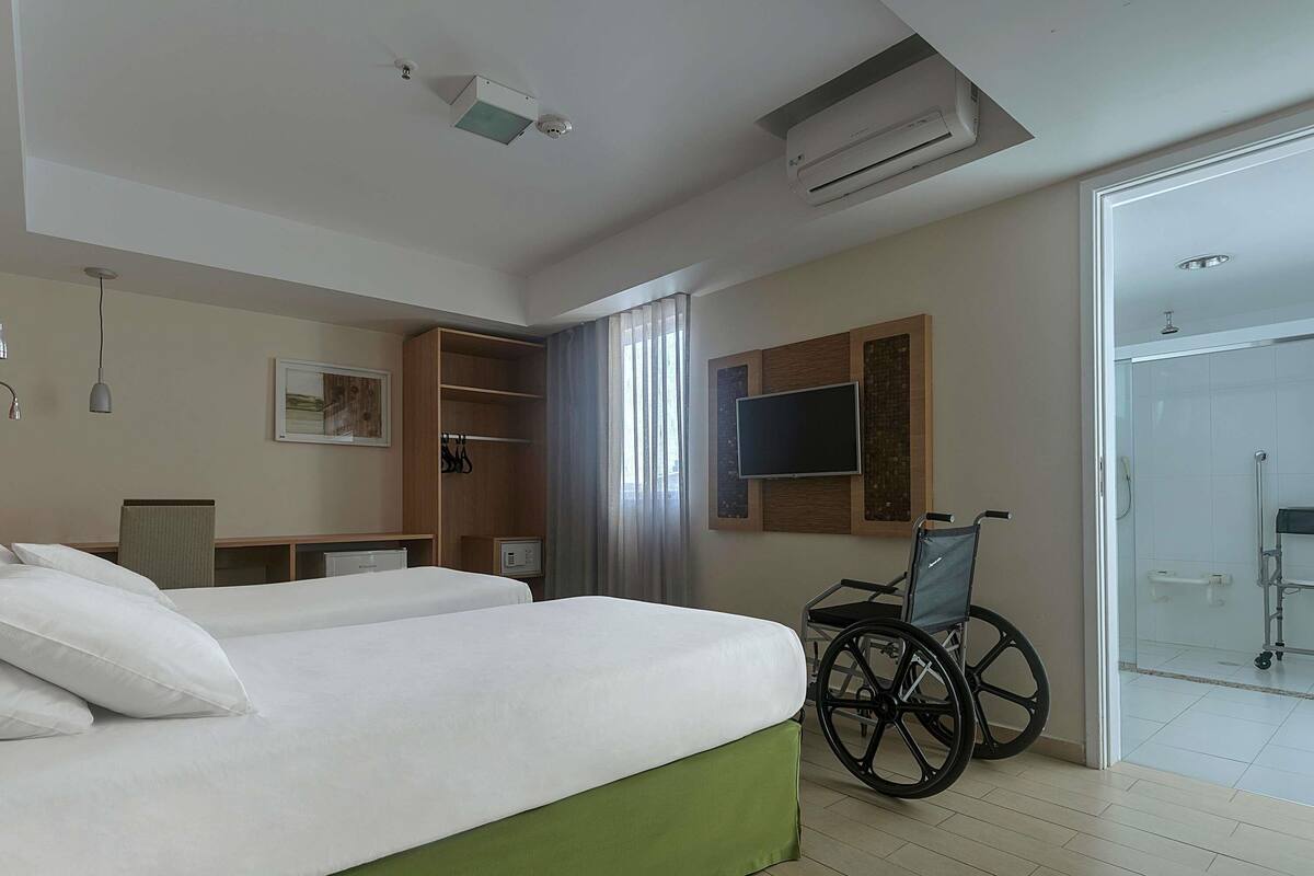 A hotel room with two beds, a wheelchair, a TV, and a shower area.