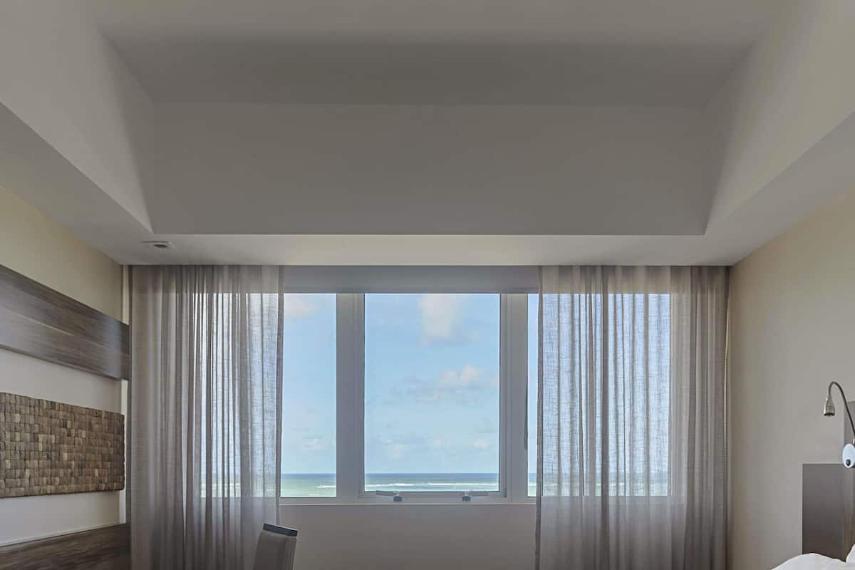 A hotel room with a large window, a bed, a chair, and a wall-mounted artwork.