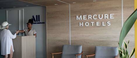 Uma receção com uma parede de madeira e um sinal a indicar 'MERCURE HOTELS'. Existem duas cadeiras cinzentas e uma planta num vaso em primeiro plano.