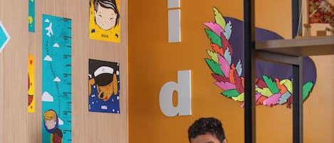 A children's play area with a height chart, a bookshelf, and a child seated at a table.