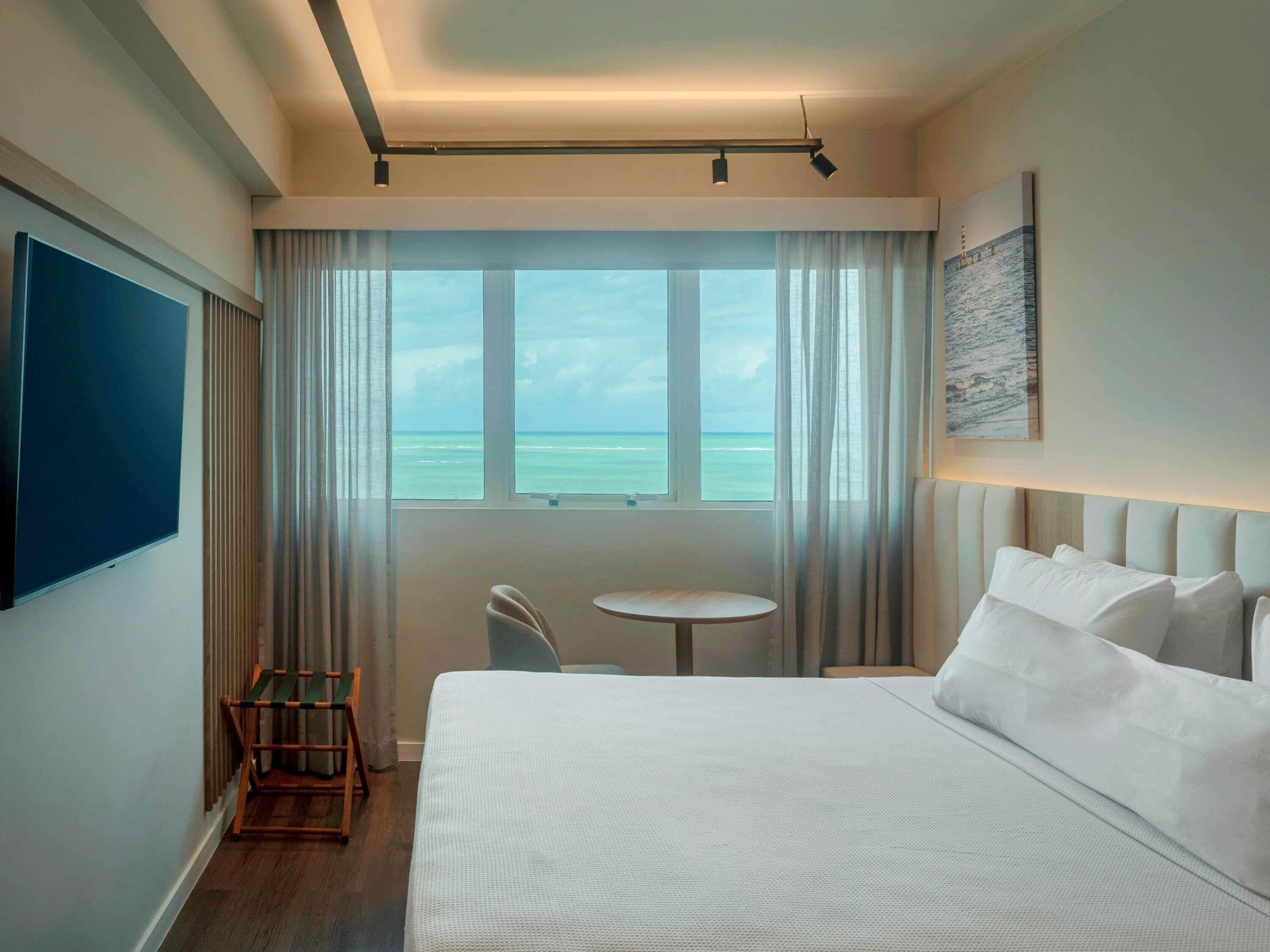 Quarto de hotel com uma cama grande, uma mesinha, uma cadeira, uma TV e uma janela com vista para o mar.