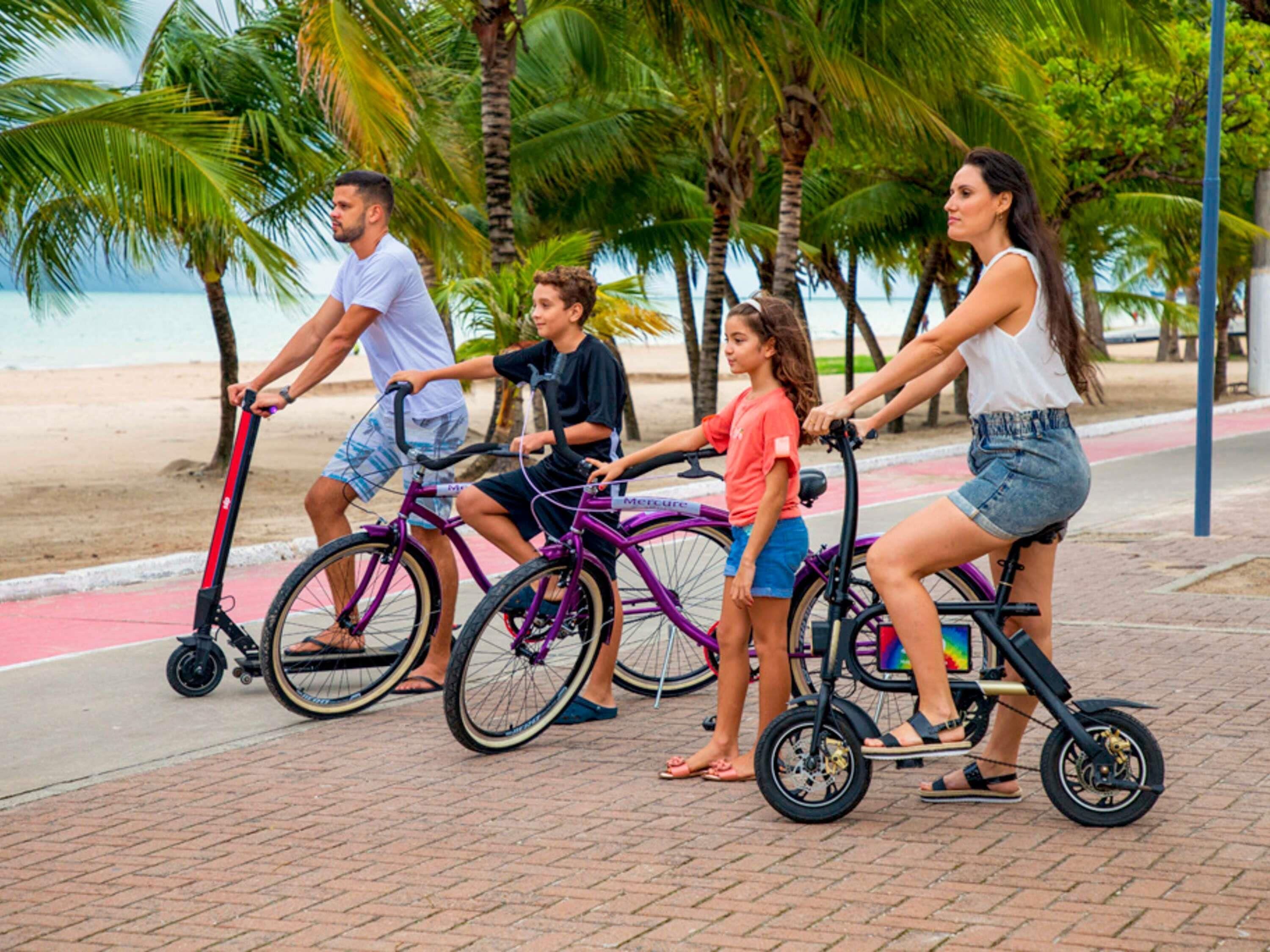 Un grupo de personas montando en bicicleta por un camino pavimentado, con palmeras y una playa al fondo.