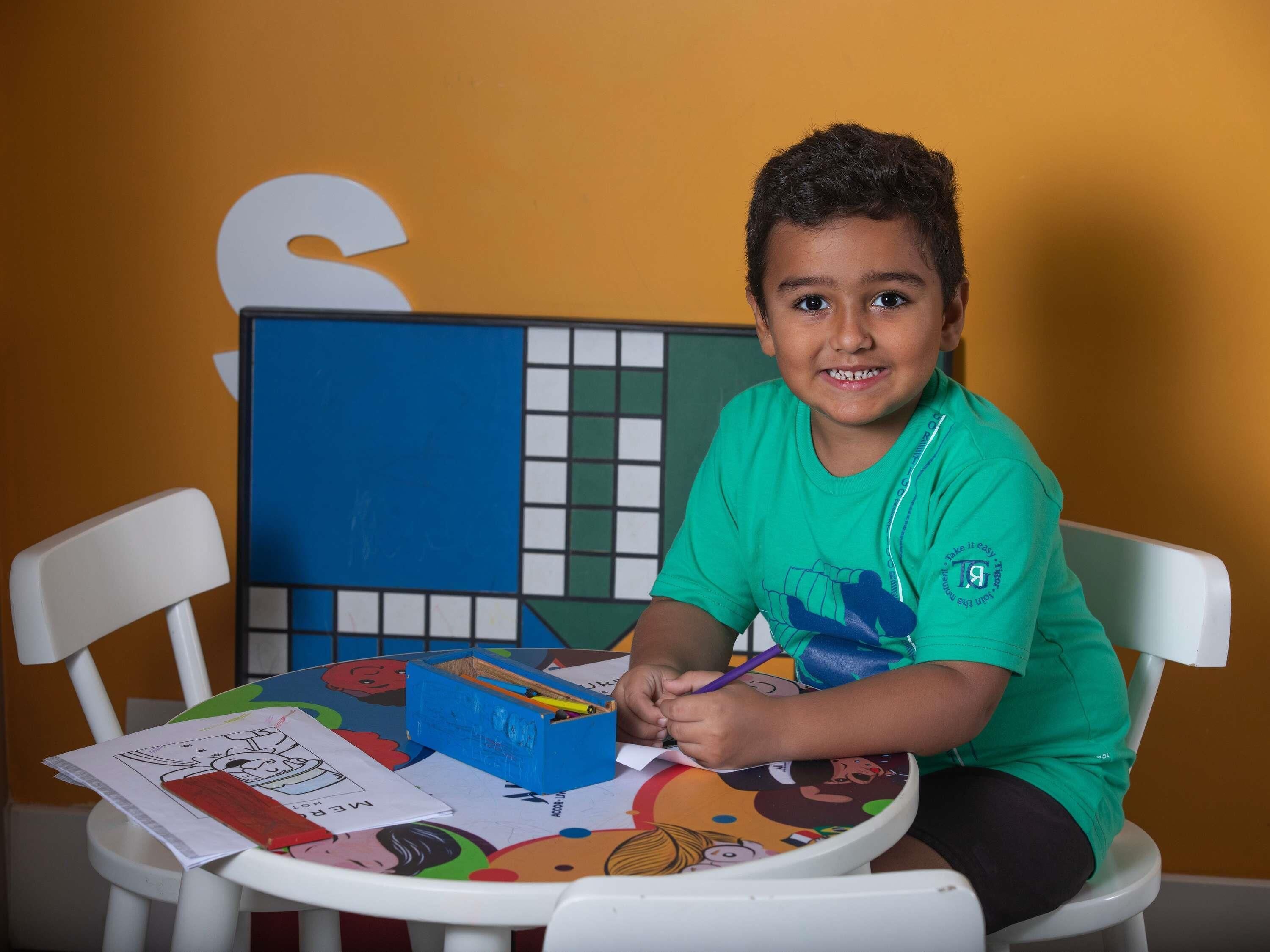 A child sitting at a table with a colorful tablecloth, a blue box, and papers.