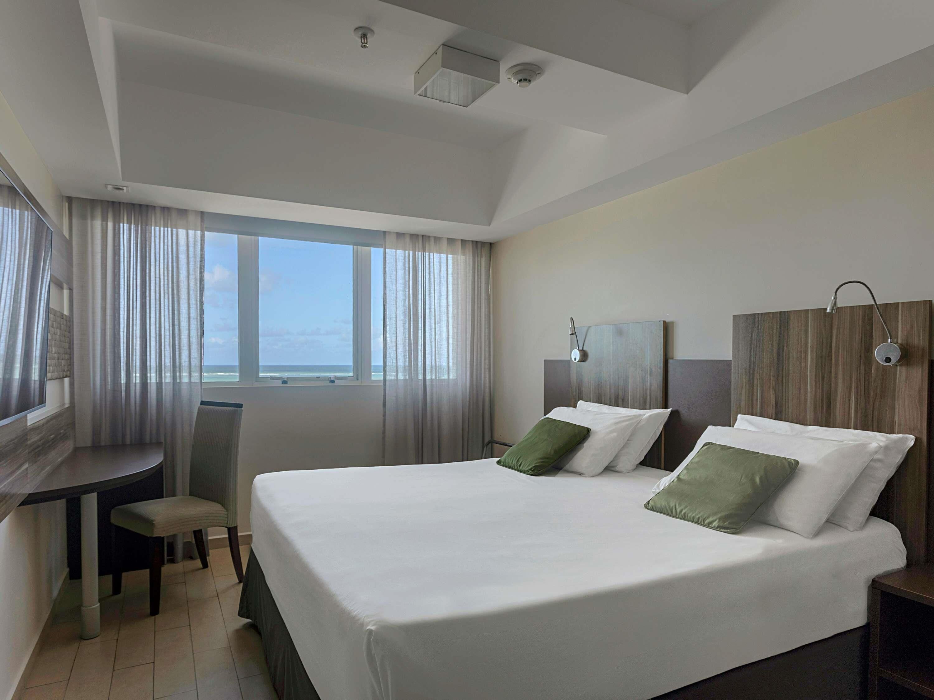 A hotel room with a large bed, two bedside tables, a desk with a chair, and a large window with curtains.