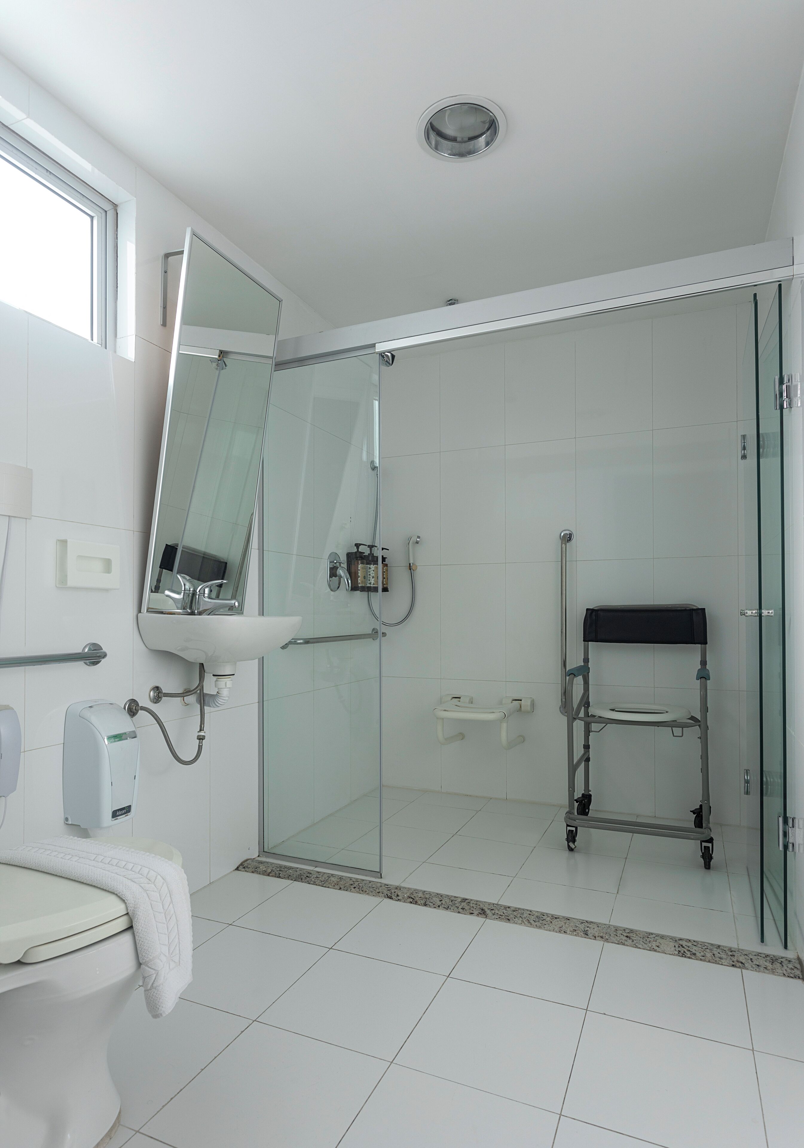 Un baño moderno con cabina de ducha de vidrio, un banco accesible para sillas de ruedas, un inodoro con toalla y un lavabo con espejo.