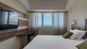 A hotel room with a large bed, a TV mounted on the wall, a desk, and a view of the ocean. - Mercure Maceio Pajucara Hotel (Maceió)