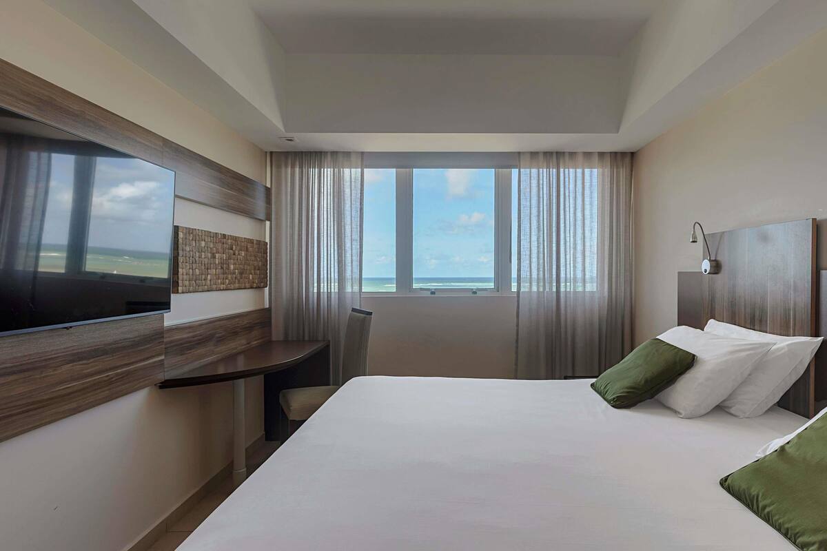 A hotel room with a large bed, a TV mounted on the wall, a desk, and a view of the ocean.