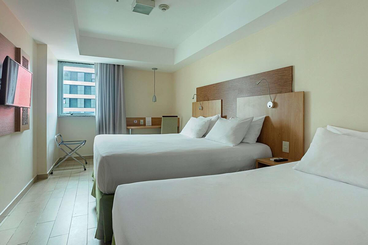 A hotel room with two beds, a desk, a chair, a window with curtains, and a ceiling light.