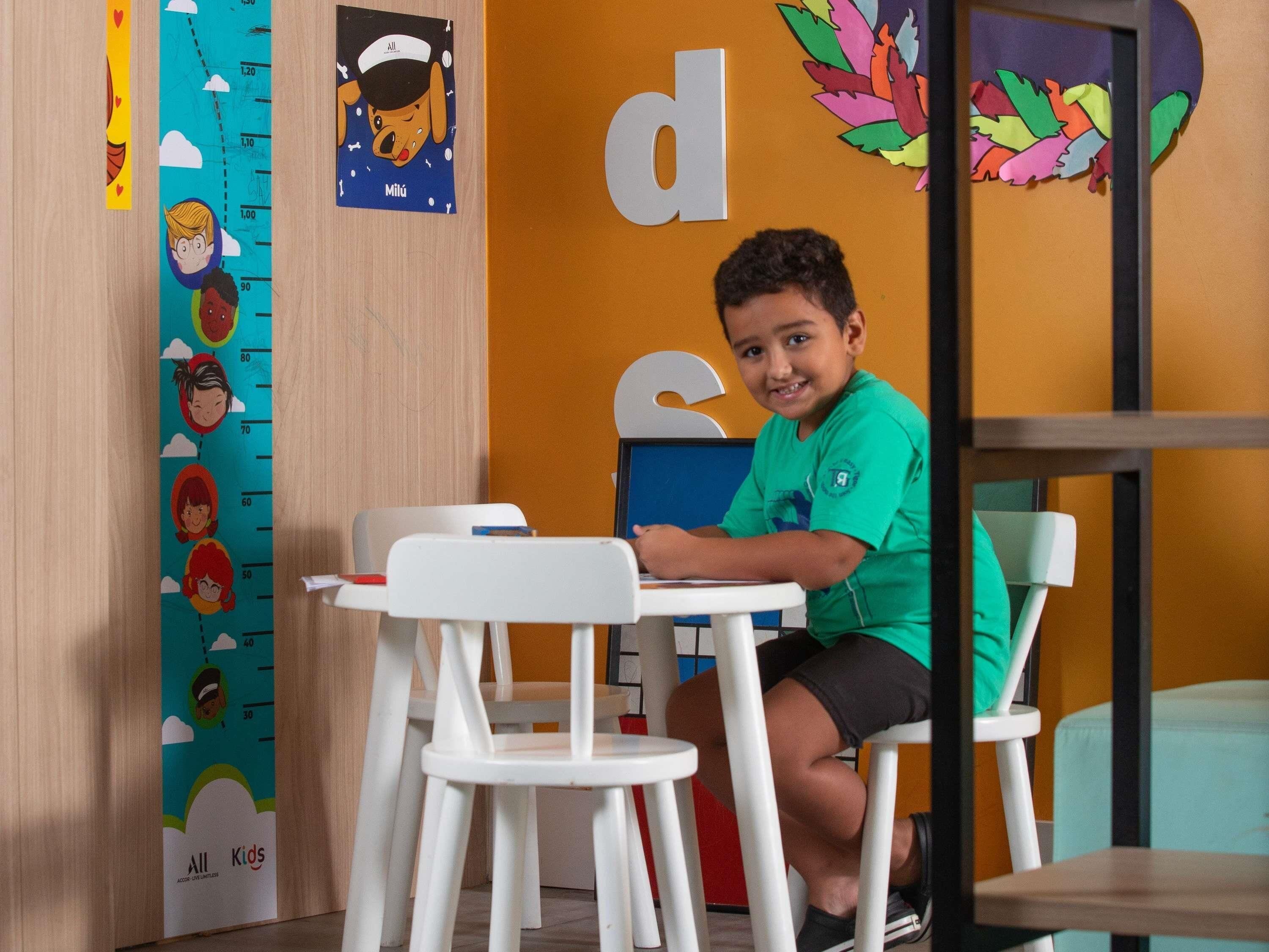 Un enfant est assis à une table blanche dans une pièce où l’on peut voir un tableau de mesure de la taille au mur et des décorations colorées.