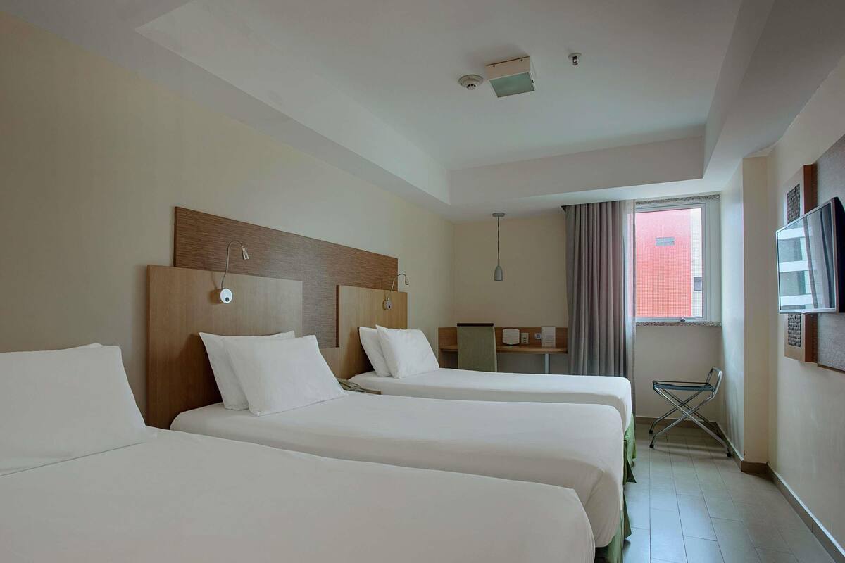 A hotel room with three beds, a desk, a chair, and a window with curtains.