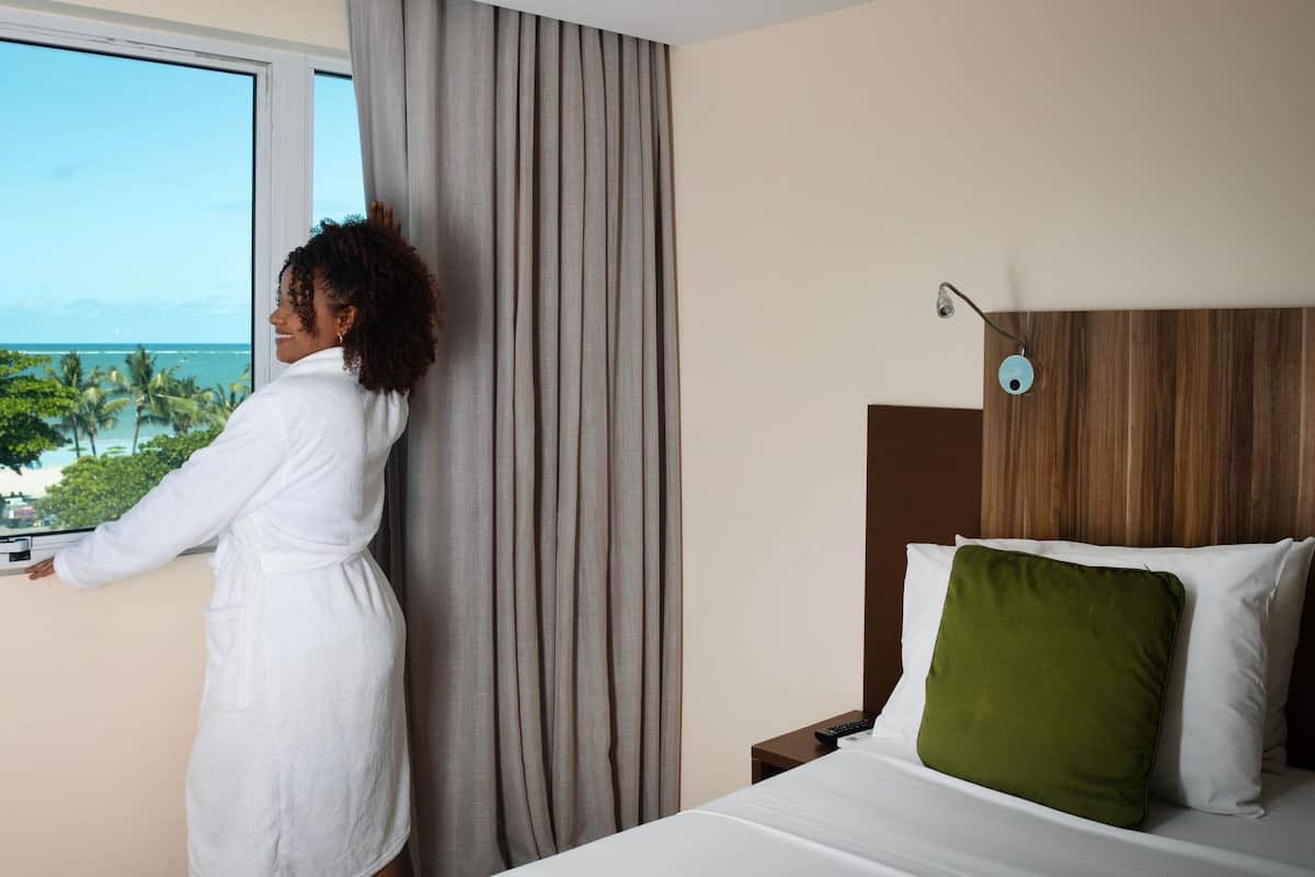 A hotel room with a bed, a nightstand, a remote control, and a view of the ocean.