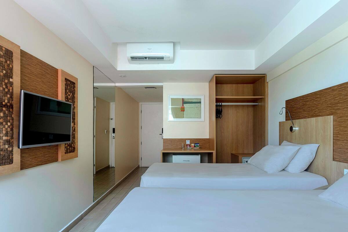 A hotel room with a bed, a TV mounted on the wall, a wardrobe, and a small desk with a chair.