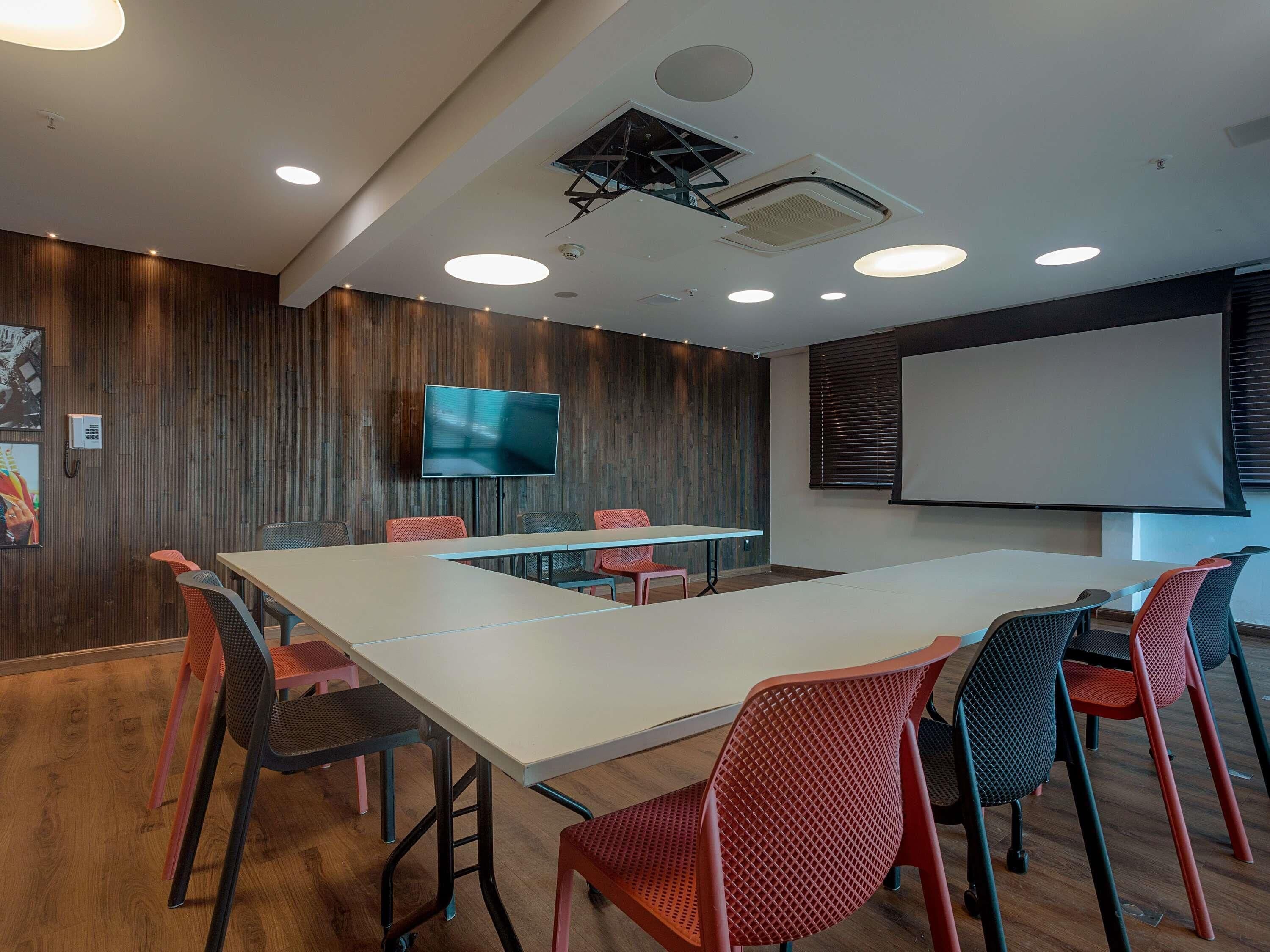 A conference room with a long table, chairs, a projector screen, and a mounted TV.