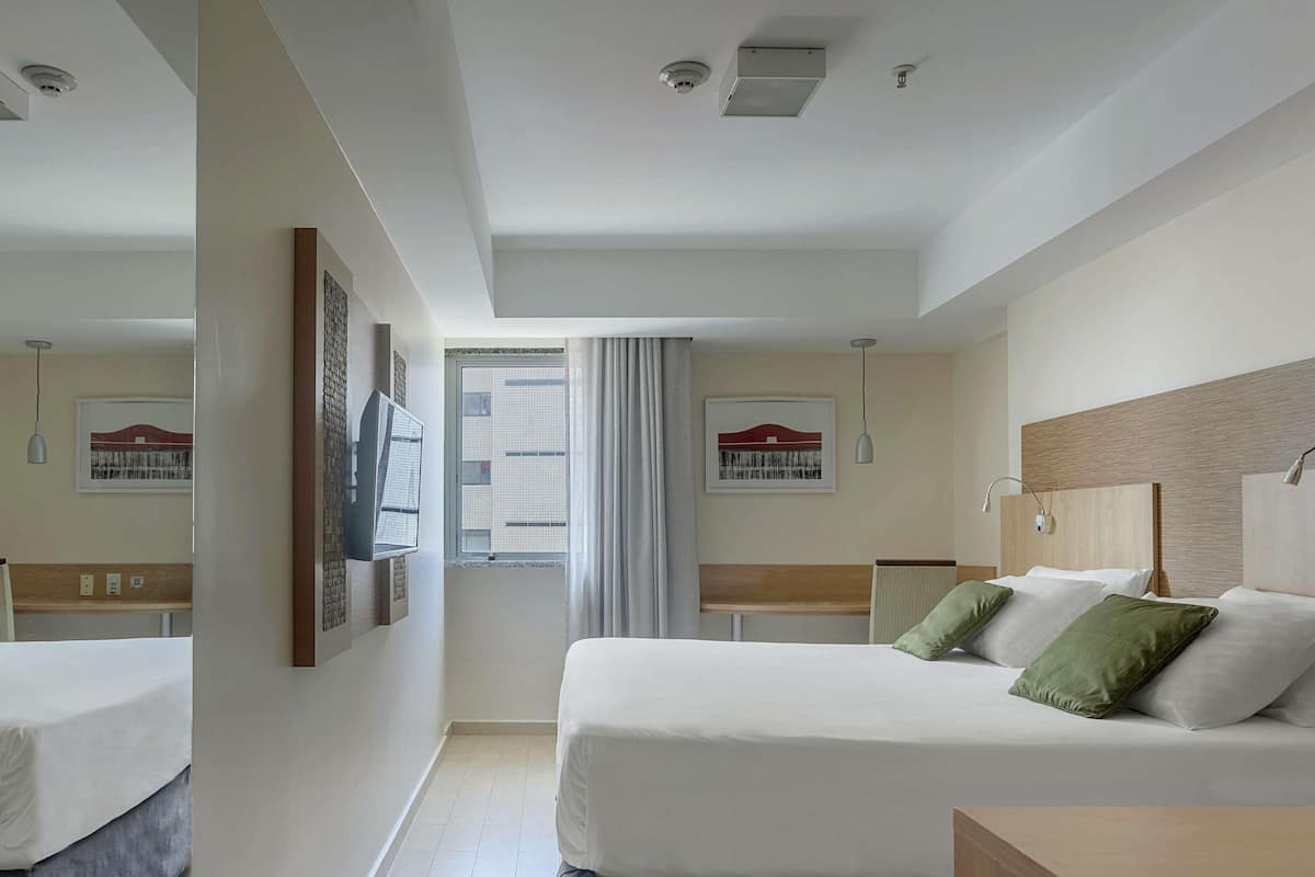 A hotel room with a bed, a bedside table, a window with curtains, and a view of a building outside.