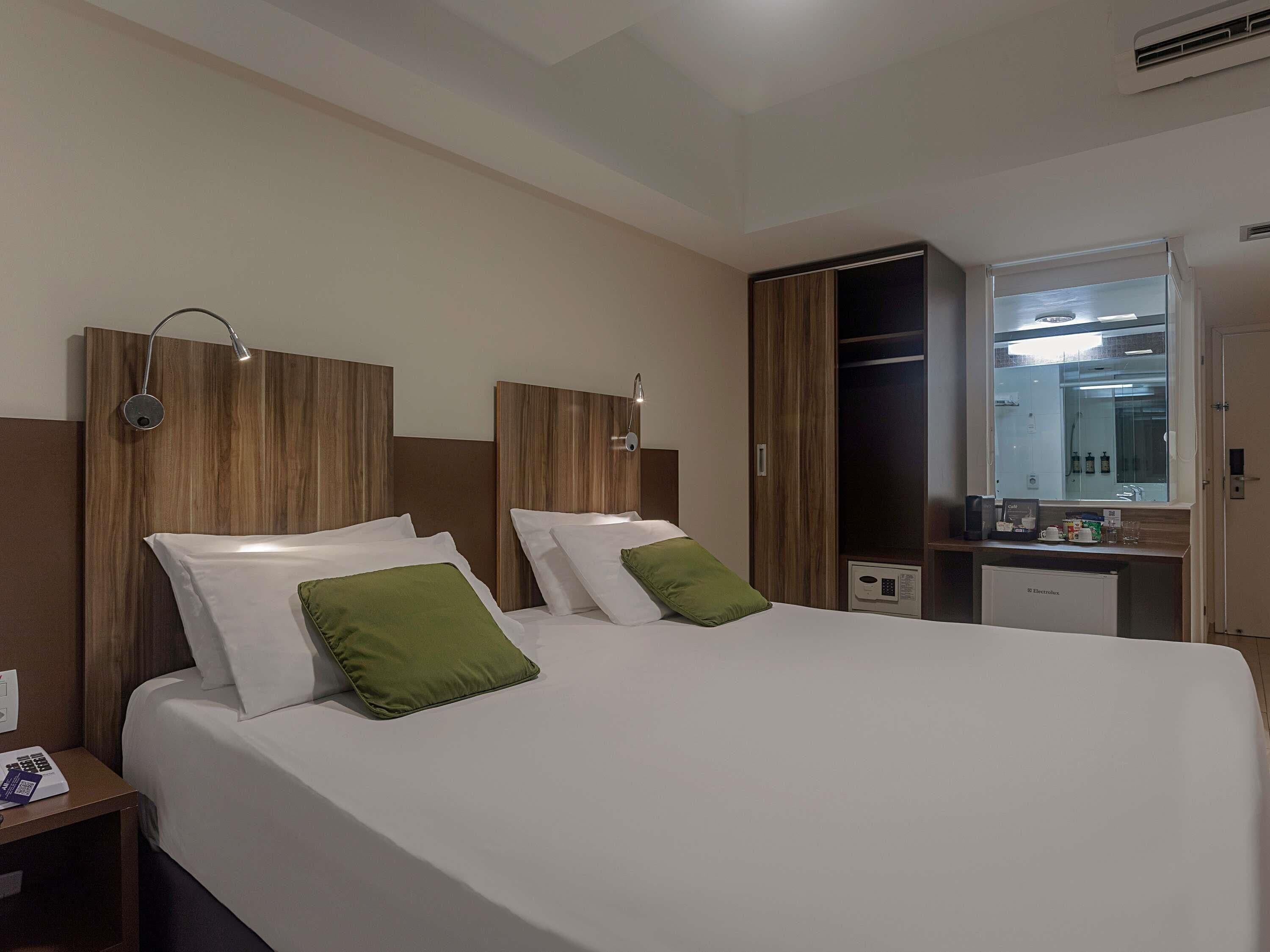 A hotel room with a large bed, two bedside tables, a wardrobe, a desk with a mirror, and an air conditioning unit.