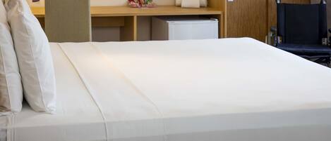 A hotel room with a bed, bedside phone, desk, chair, and a small refrigerator.