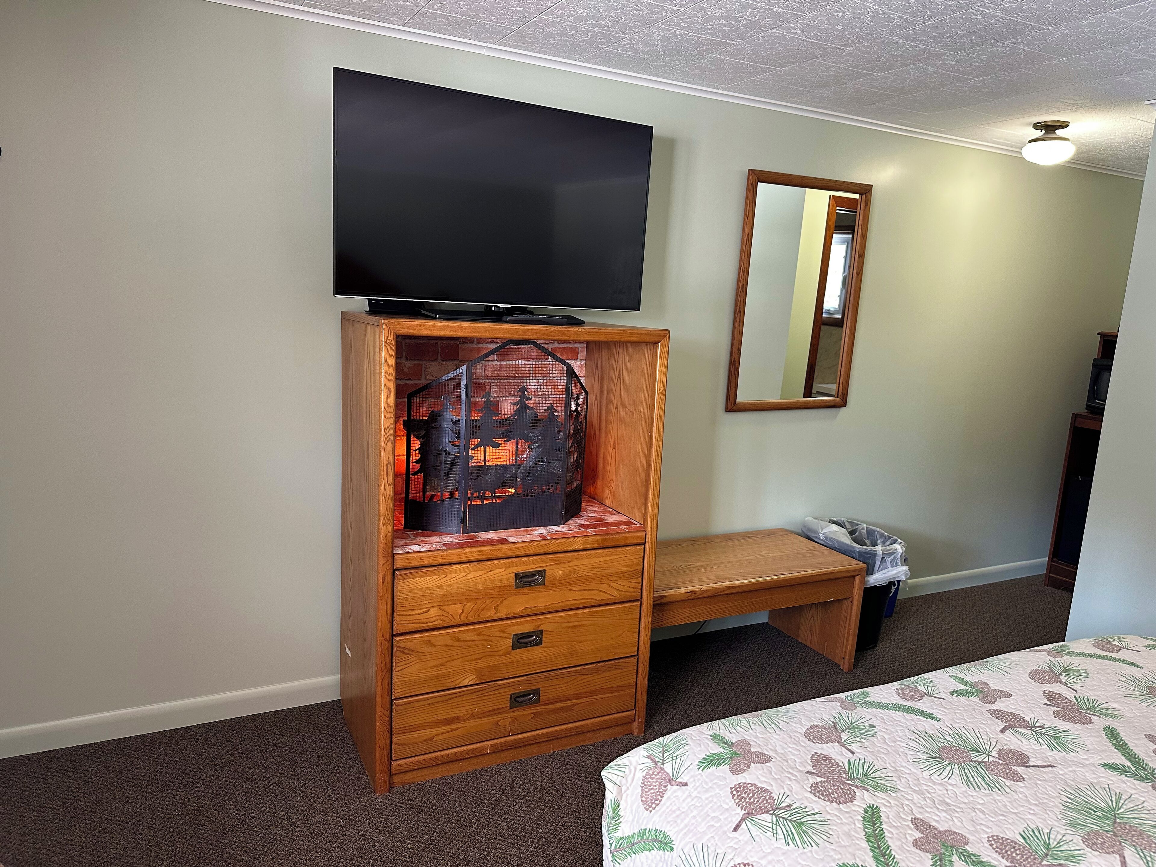A bedroom with a wooden dresser, a flat-screen TV, a bench, a bed with a pine branch bedspread, and a mirror on the wall.