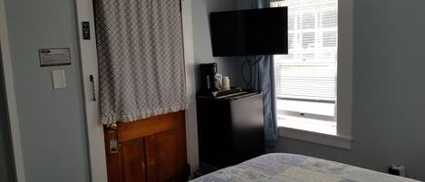 A bedroom with a bed, a television mounted on the wall, a coffee maker, and a window with blinds.