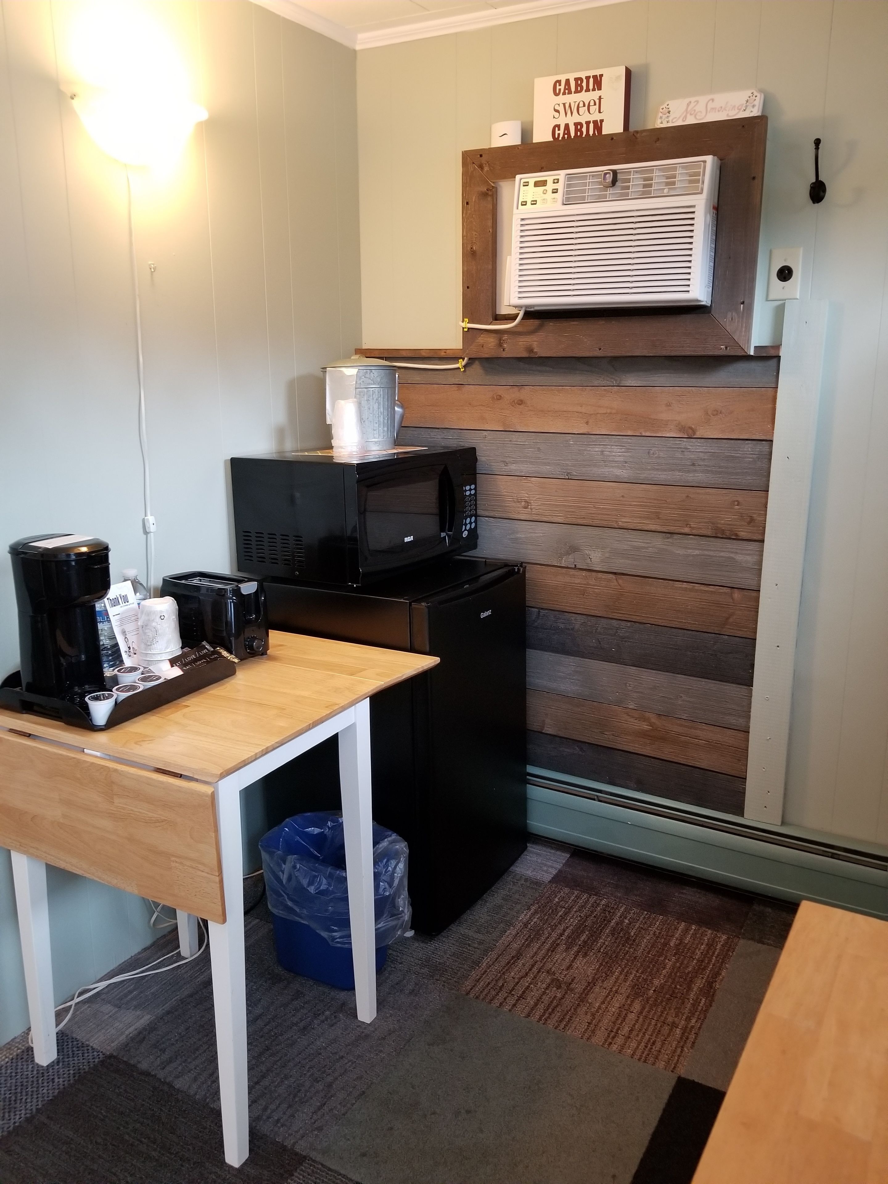 A compact kitchenette with a microwave, toaster, and coffee maker on a small table. A refrigerator is visible in the background, and a sign on the wall reads "CABIN SWEET CABIN."