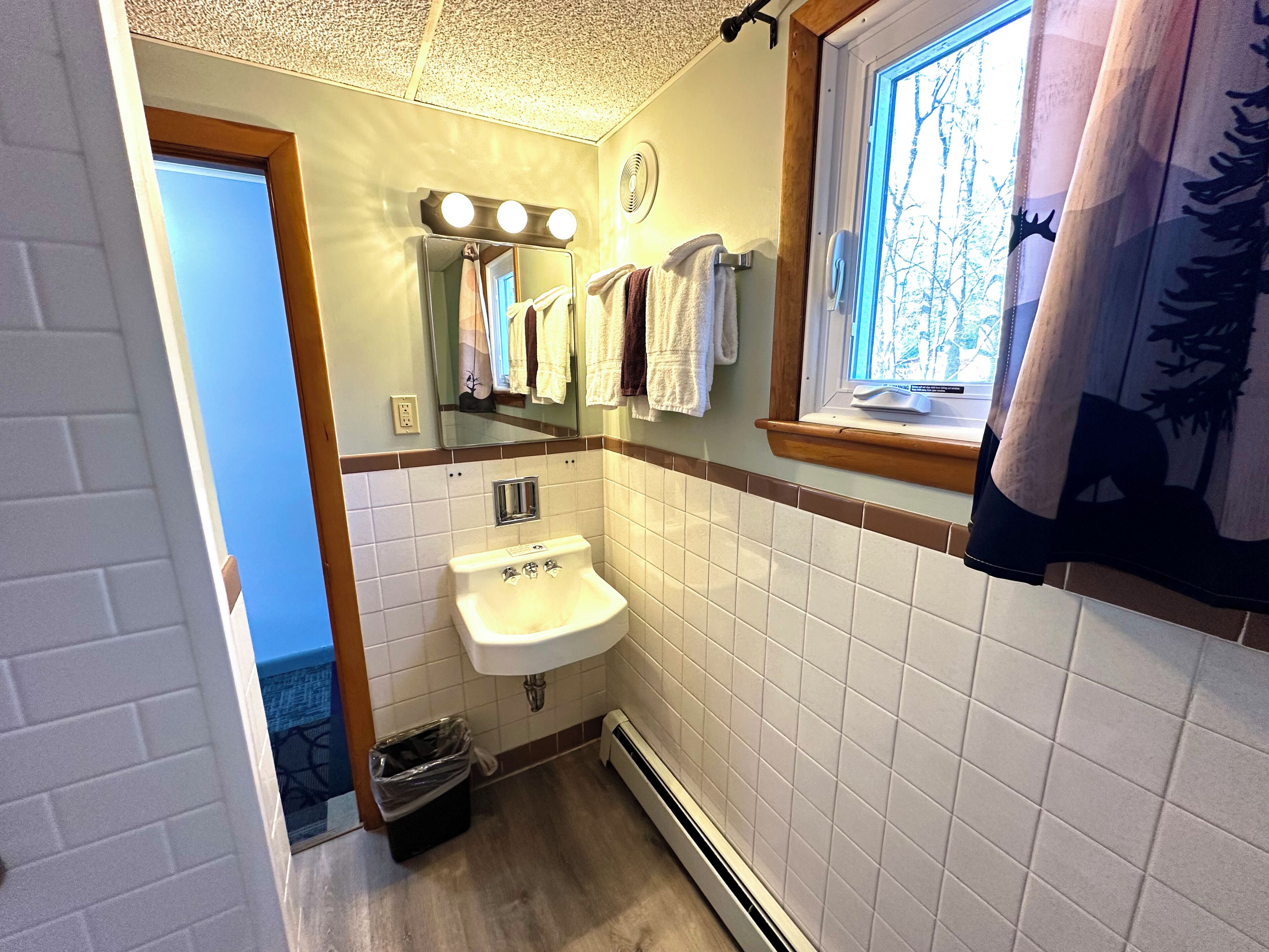 A bathroom with a sink, mirror, and window. There are towels hanging on the wall and a trash can.