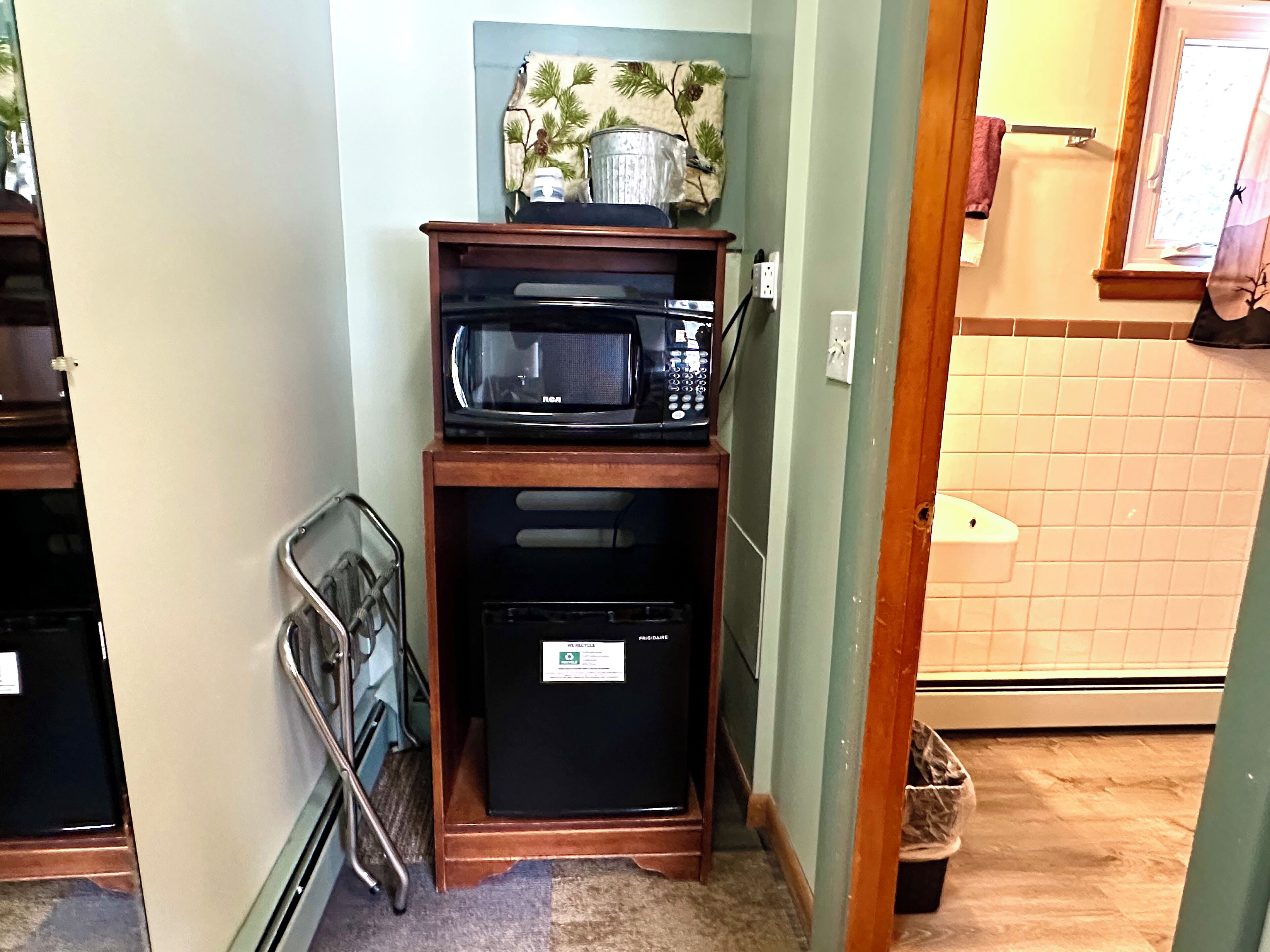 Cocina pequeña con microondas y un refrigerador compacto sobre una repisa de madera, una silla metálica y un cesto de basura.