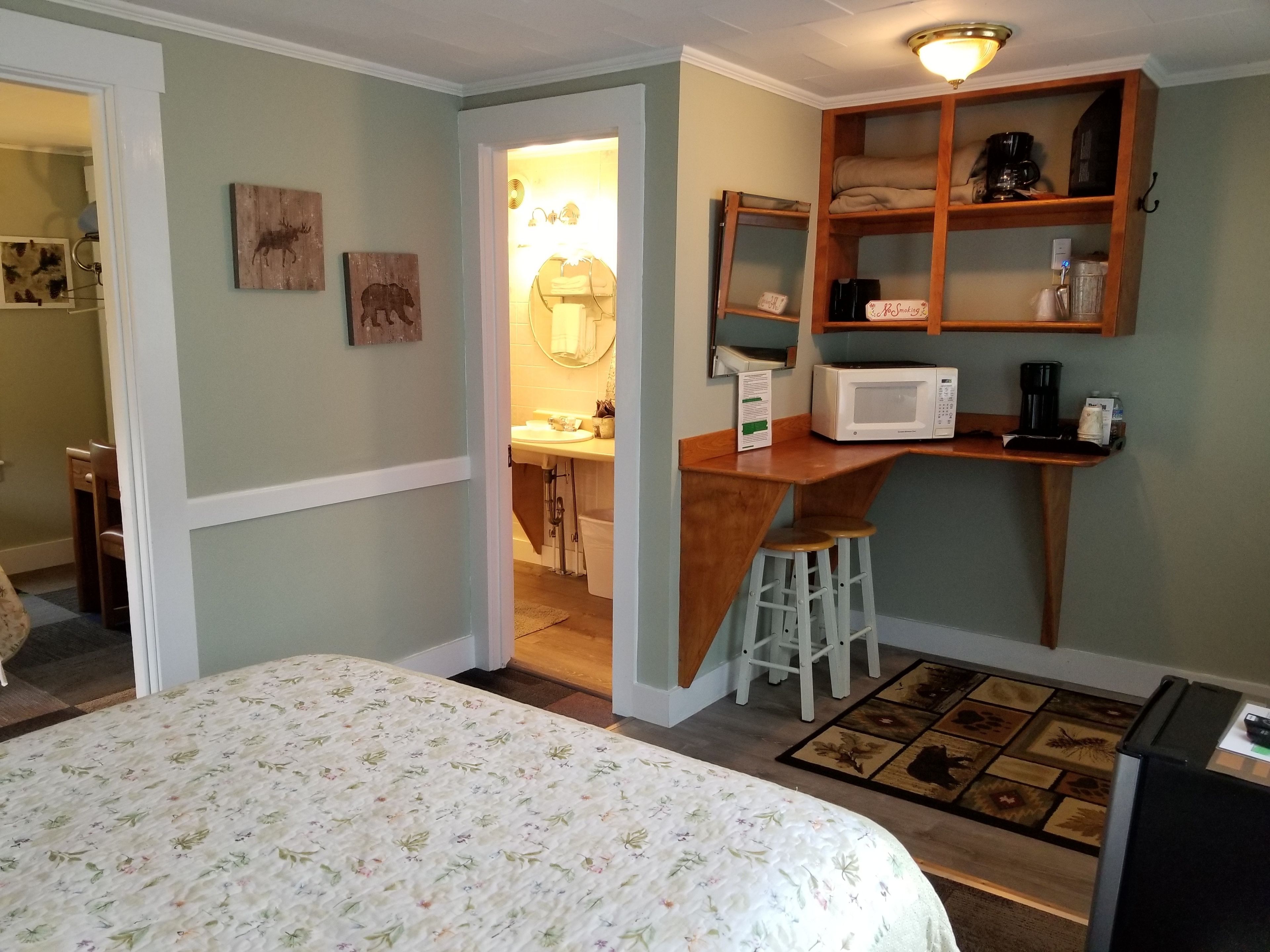 Une pièce compacte comprenant un lit, une petite kitchenette, une étagère en bois avec un four à micro-ondes, et une salle de bain visible à travers une porte ouverte.
