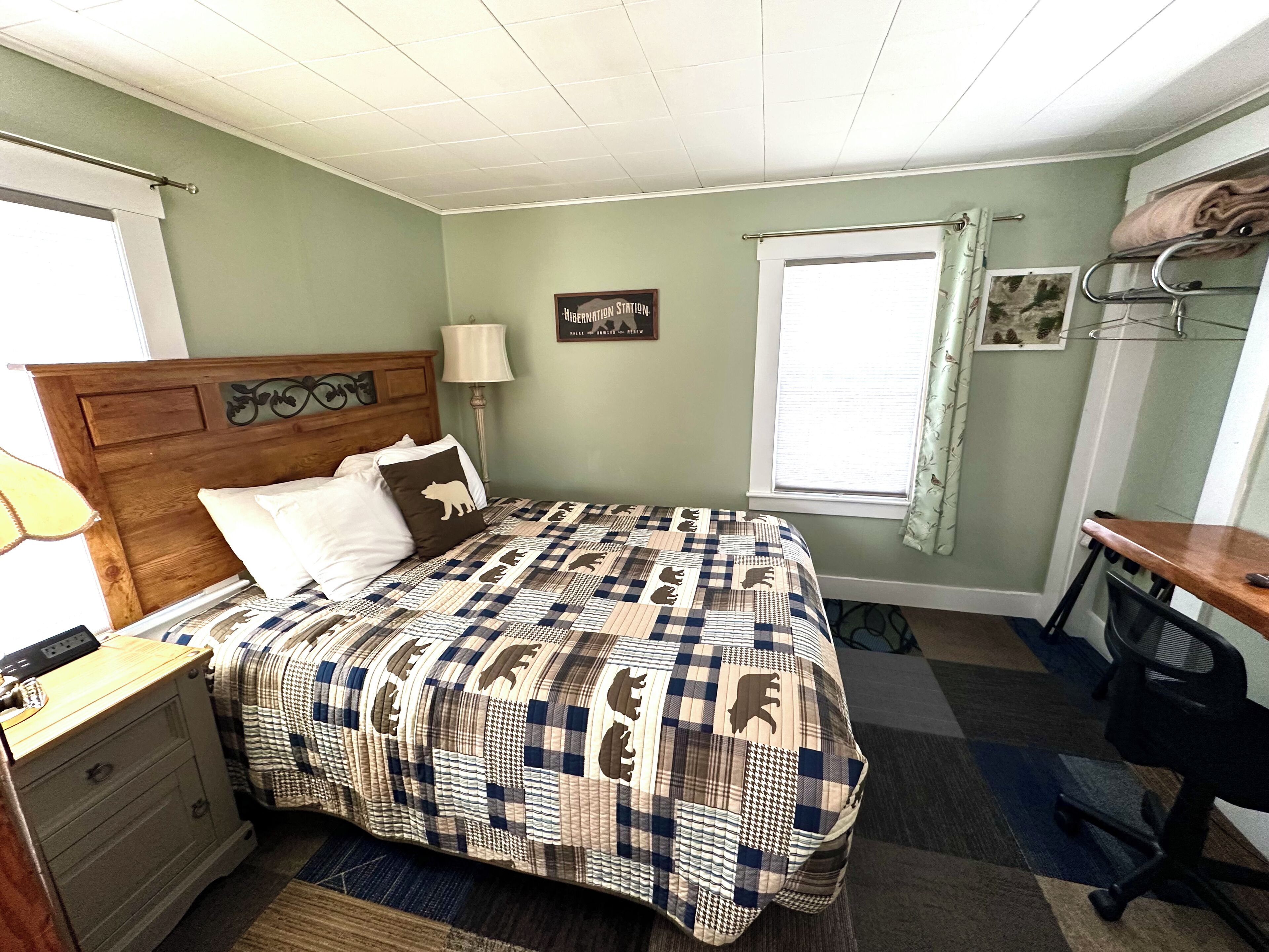 A bedroom with a bed, a nightstand, a desk, a lamp, a window with curtains, and a wall decoration.