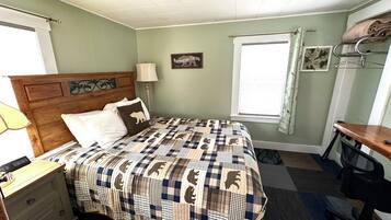 A bedroom with a bed, a nightstand, a desk, a lamp, a window with curtains, and a wall decoration.