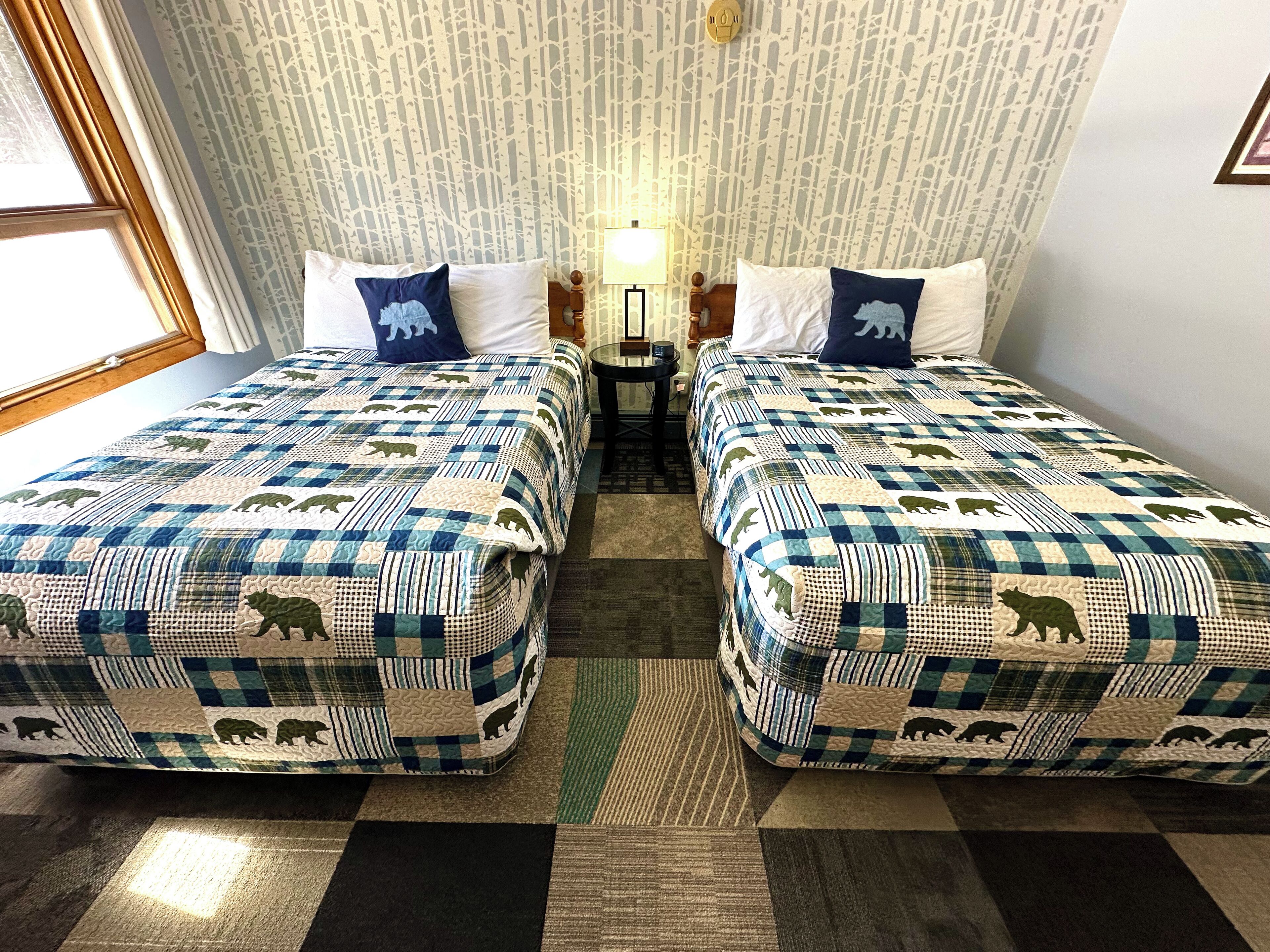 Ein Hotelzimmer mit zwei Betten, jeweils mit einer blau-weiß karierten Tagesdecke mit Bärenmotiven. Zwischen den Betten steht ein kleiner Tisch, an der Wand hängt eine Lampe und es gibt ein Fenster mit Vorhängen.