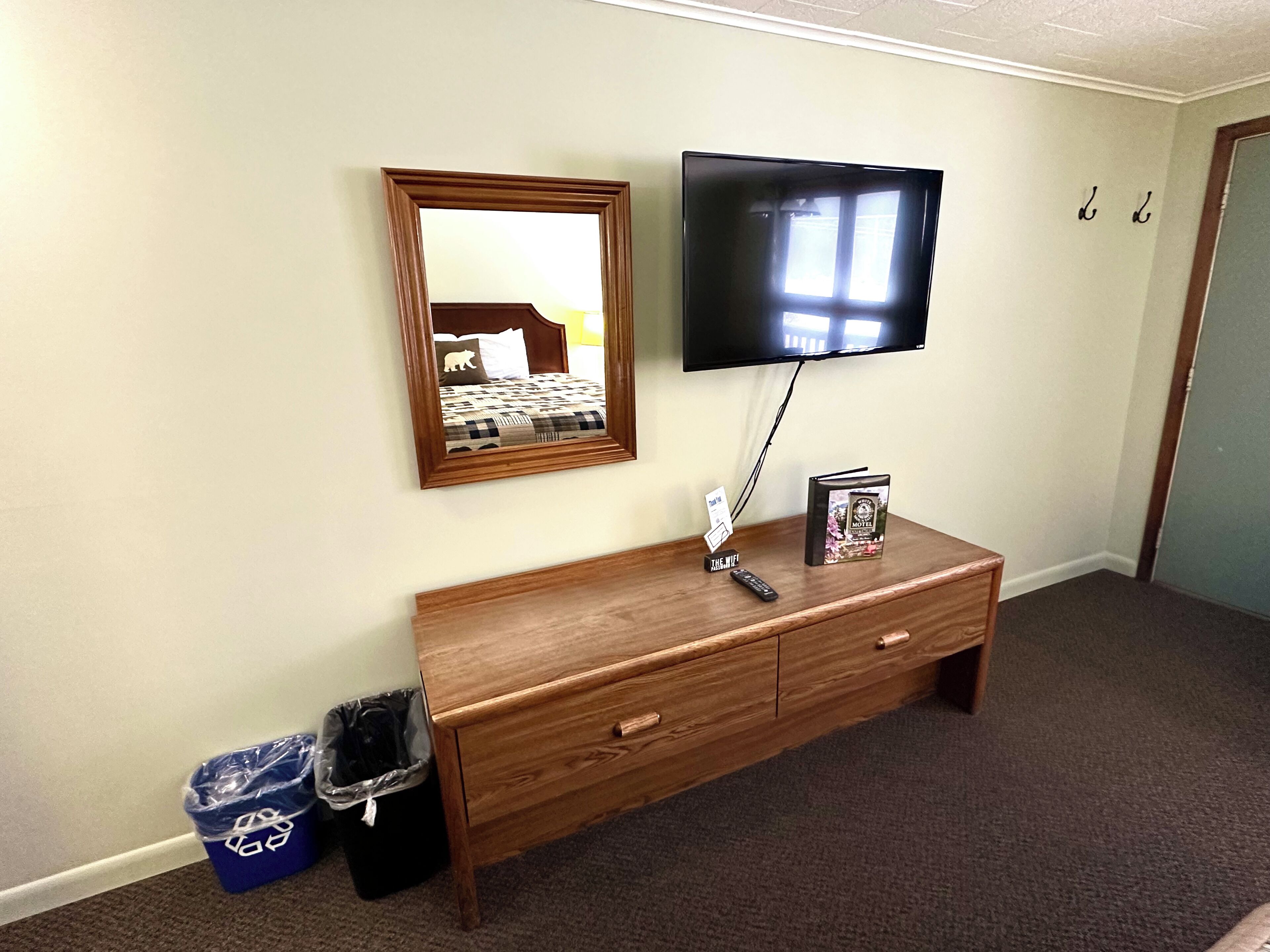 A hotel room with a wooden dresser, a flat-screen TV mounted on the wall, a mirror, and a trash bin.