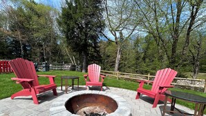 Un brasero rodeado de sillas Adirondack rojas y pequeñas mesas, ubicado en un patio empedrado con borde de piedra.
