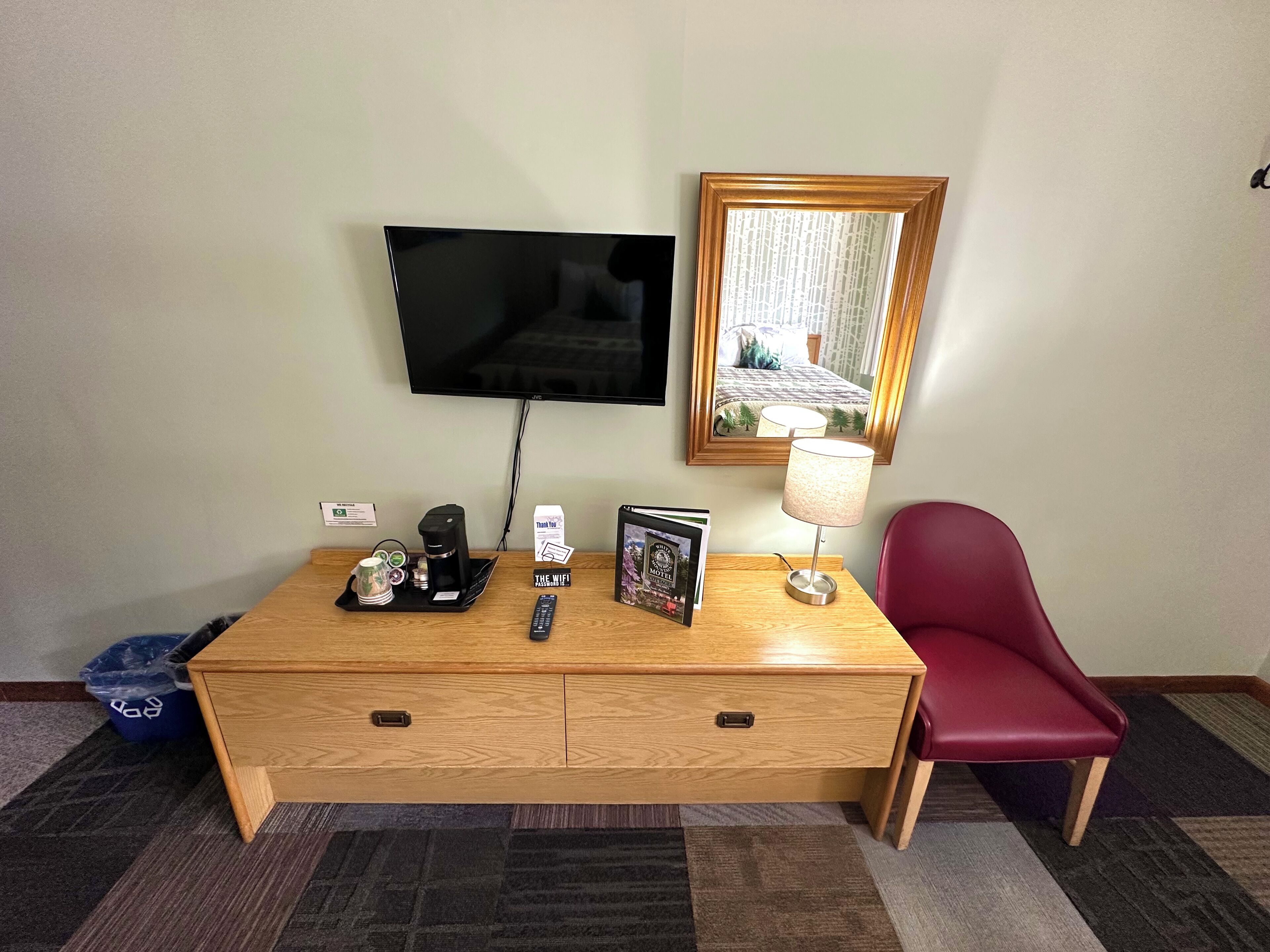 A hotel room with a wooden desk, a television, a mirror, a red chair, a lamp, and a tray with a coffee maker and a remote control.
