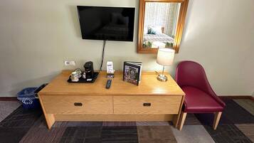 A hotel room with a wooden desk, a television, a mirror, a red chair, a lamp, and a tray with a coffee maker and a remote control.